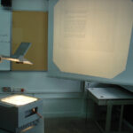 An overhead projector in the corner of a dingy classroom lighting up a pull-down screen.