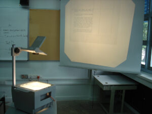 An overhead projector in the corner of a dingy classroom lighting up a pull-down screen.