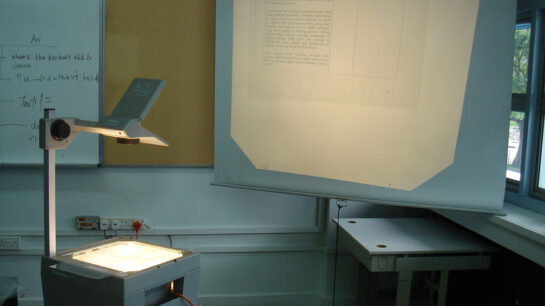 An overhead projector in the corner of a dingy classroom lighting up a pull-down screen.