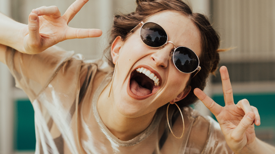Laughing woman in dark glasses showing victory sign in both hands.