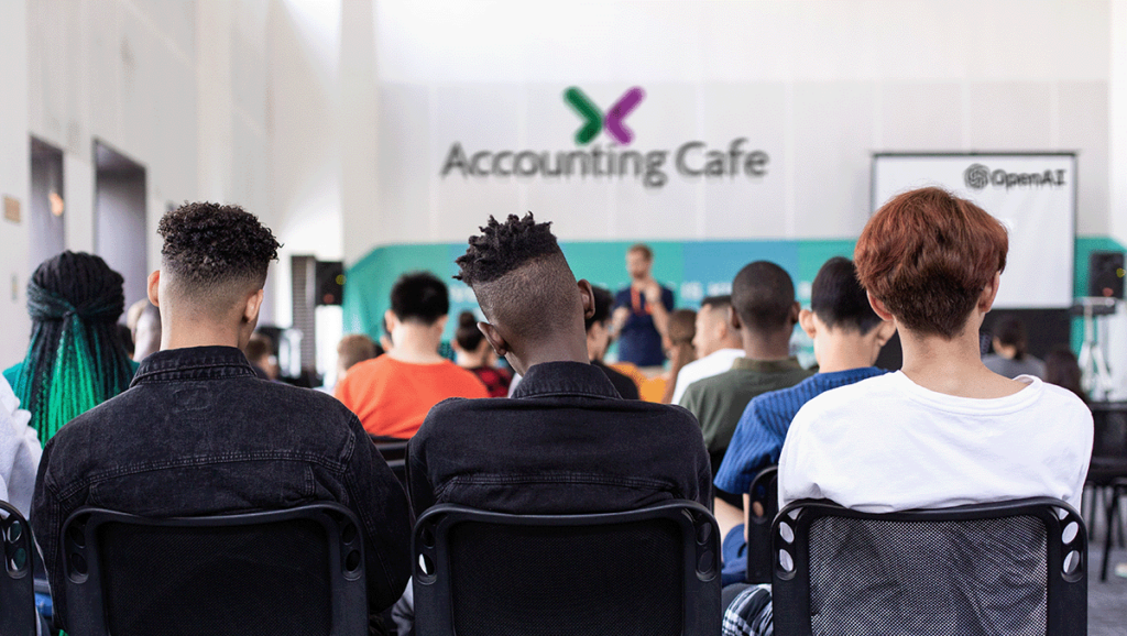 Photograph from the back of a class full of students. The room has the Accounting Cafe logo on the wall and "Open AI" on a digital whiteboard.
